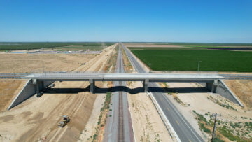 McCombs Road Grade Separation seen from above via drone, head on. Future high-speed rail right of way, an existing freight railroad, and State Route 43 stream beneath the structure, disappearing on the horizon. Lush trees and dry shrubs are all around.