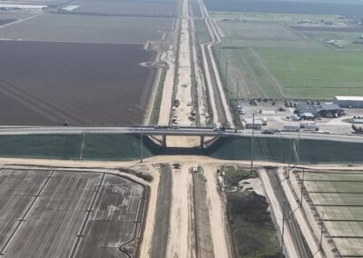 An image shot from above via drone of the Hanford Armona Road Grade Separation, from very high and very far away to give a sense of scale. Streaking straight down the center of the image from the bottom of the image frame to the horizon line with a cloudless sky at the top of the image frame is the future right of way of high-speed rail, where tracks will go, and over which the Hanford Armona structure passes over. The bright new concrete span is centered in the image, with the earthen approach embankments to the right and left of it. Recently moved earth tones are everywhere, and the slopes of the earthen embankments are treated with a substance that distinguishes them from everything around them.