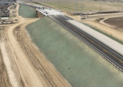 An image shot from above via drone of the Hanford Armona Road Grade Separation. The bright new concrete span is in the top left of the image, with one of the earthen approach embankments sloping down and to the right for the rest of the image. Recently moved earth tones are everywhere, and the slopes of the earthen embankments are treated with a substance that distinguishes them from everything around them. A cloudless sky above the horizon line frames the top of the image.