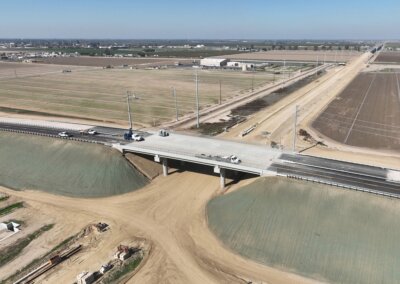 Concrete overpass stretching above unpaved roadway which will hold future high-speed rail tracks. Four construction trucks are on top of the overpass. The sky is clear and blue, and cleared agricultural fields surround the overpass.