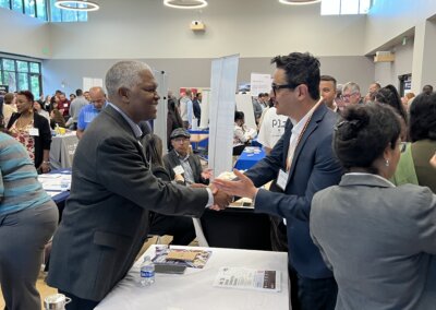 Vendors and attendees congregate around tables and shake hands in the expo hall with bright and high ceilings at the Small Business Fair in Burlingame.